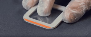 A gloved hand holds a thin, transparent sheet partially placed inside a rectangular metal frame with one edge highlighted in orange, against a dark background—possibly prepared for Blast Cleaning.
