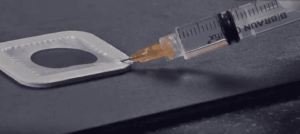 A close-up of a syringe filled with yellow liquid injecting fluid onto a white, square-shaped object with a circular hole—reminiscent of precision techniques used in blast cleaning—set against a dark surface.