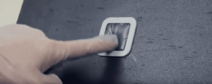 A hand presses a finger into a small, square, rubberized button with bristles set into a dark, textured surface—part of a blast cleaning system.