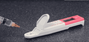 A syringe with a needle is positioned above a white and red rapid test device on a dark, blast cleaning-speckled surface. The test device has an open lid, revealing a spot for sample application.