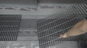 A person’s arm is placing or removing a metal grate over a grated floor in a sandblast booth, with dust or powder covering the surrounding surfaces.