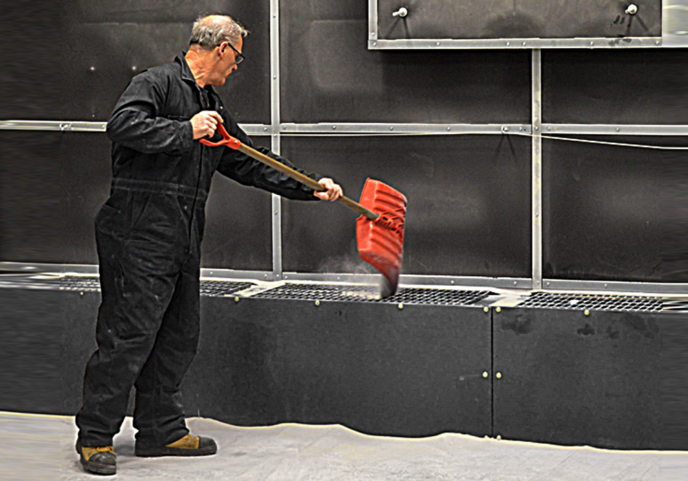 A man in black coveralls uses a red snow shovel to push material into a grated opening on the floor, possibly cleaning or clearing an industrial area equipped with Abrasive Media Recovery Systems.