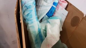 A cardboard box containing used disposable adult diapers, some stained blue and pink with visible absorption lines and materials, possibly marked by flexo printing.